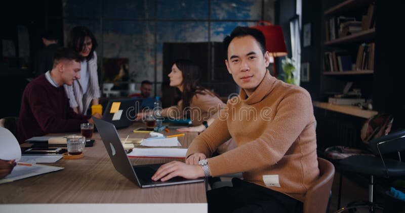 En bild av en lycklig ung affärsman som leker i kameran med hjälp av en bärbar dator som står på en modern, loft office-arbetsplat