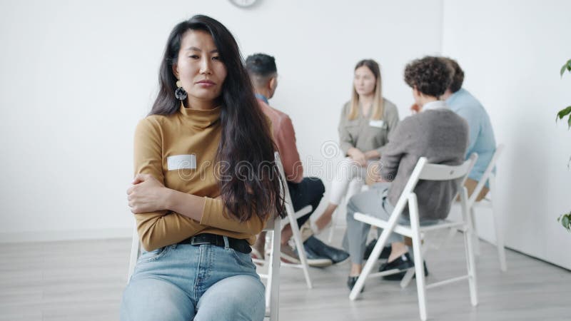 En bild av stressad ung asiatisk flicka som tittar på kameran medan människor i bakgrunden talar vid gruppterapimöte