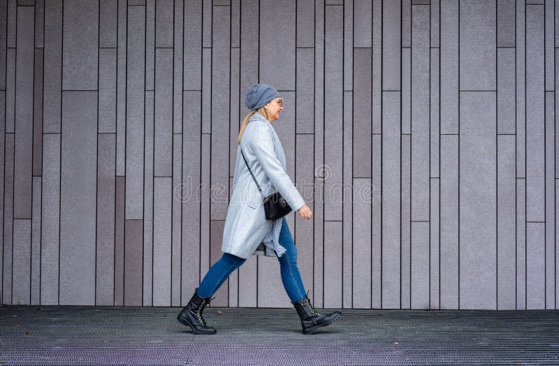 En blond kvinna i grå kappjejeans vinterhatt och läderhandväska som går mot bakgrund av grå modern vägg arkivbilder