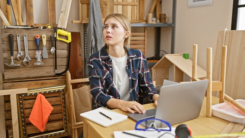En blond kvinna med blå ögon överväger ett projekt i en konstgjord snickerverkstad med verktyg och trä när man arbetar på en bärbar dator