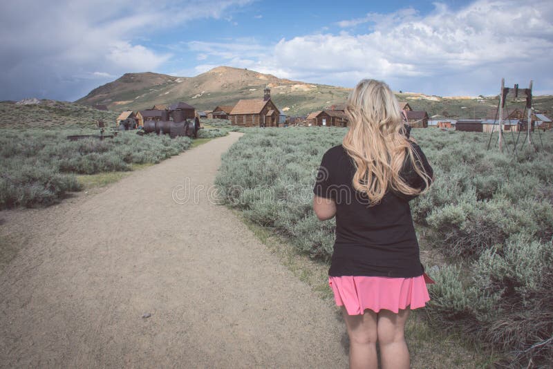En blond kvinnlig går på en bana in mot mer övergiven byggnader i spökstaden av Bodie California royaltyfri fotografi En blond kvinnlig går på en bana in mot mer övergiven byggnader i spökstaden av Bodie California royaltyfri fotografi