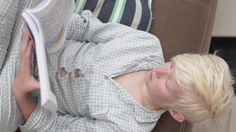 En blond pojke i pyjamas som läser en bok hemma på soffan på morgonen. arkivfilmer