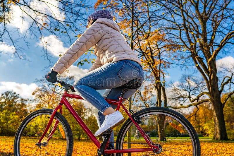 En blondinerkvinna med beige jejans och vinterhatt cyklar i stadsparken i autumnalundervisning på soliga dagar. arkivfoto