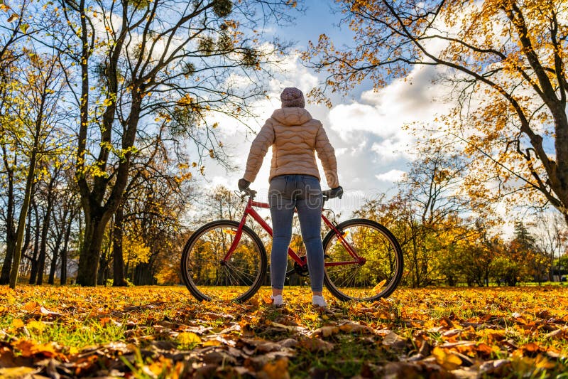 En blondinerkvinna med beige jejans och vinterhatt cyklar i stadsparken i autumnalundervisning på soliga dagar. royaltyfri fotografi