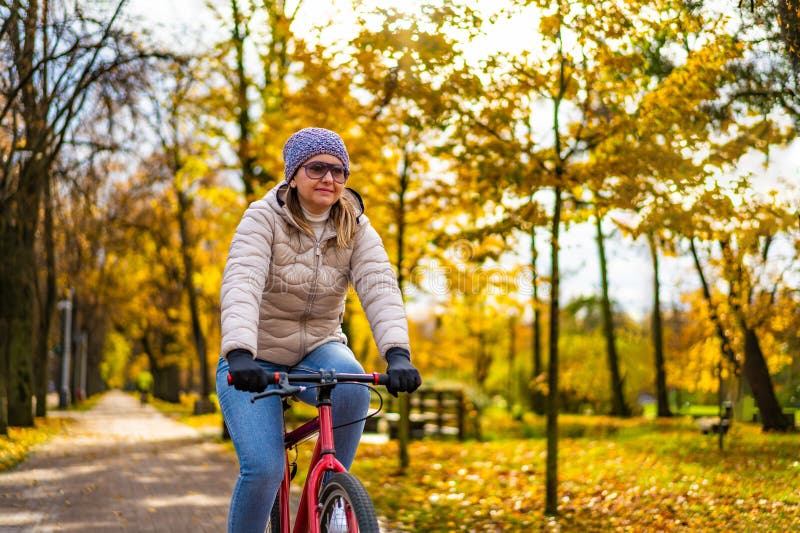 En blondinsk kvinna med beige jejans och vinterhatt cyklar i stadsparken i autumnalundervisning på soliga dagar arkivfoto
