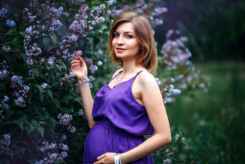 En charmig härlig ung gravid kvinna i purpurfärgad violett klänning i en blommande lila trädgård ser kameran med mjukhet fotografering för bildbyråer En charmig härlig ung gravid kvinna i purpurfärgad violett klänning i en blommande lila trädgård ser kameran med mjukhet fotografering för bildbyråer