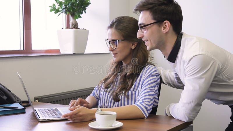 En el hombre y la mujer de la oficina con los vidrios que sonríen en el escritorio delante del ordenador portátil