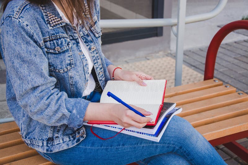 En flicka i jeanskläder sitter på en bänk, rymmer anteckningsböcker och skriver På gatan skriver hon i en anteckningsbok som stud arkivfoto En flicka i jeanskläder sitter på en bänk, rymmer anteckningsböcker och skriver På gatan skriver hon i en anteckningsbok som stud arkivfoto