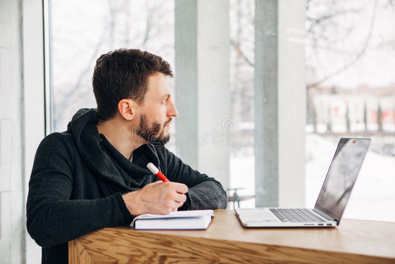 En freelancer skriver en anmärkning i en anteckningsbok och ser ut fönstret arkivbilder En freelancer skriver en anmärkning i en anteckningsbok och ser ut fönstret arkivbilder