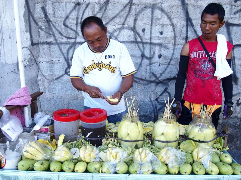 En fruktförsäljare som skivar en grön mango, medan en kund ser på royaltyfria foton En fruktförsäljare som skivar en grön mango, medan en kund ser på royaltyfria foton