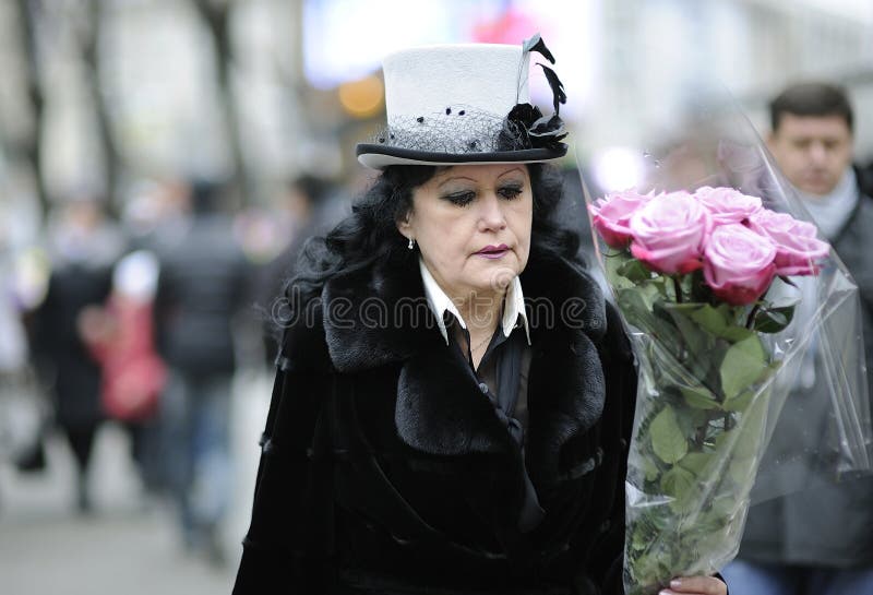 Old woman wearing retro style hat and coat walking down the street with a bouquet of roses in hand, blurred people on a background. January 9, 2012. Kyiv, Ukraine. Old woman wearing retro style hat and coat walking down the street with a bouquet of roses in hand, blurred people on a background. January 9, 2012. Kyiv, Ukraine