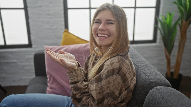 En glad ung kaukasisk kvinna med utsträckta handflator på en soffa inomhus som log lätt arkivbild