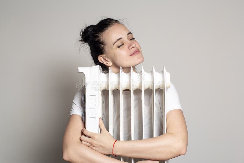 En glad, ung kvinna kramar en elektrisk radiator och njuter av värmen, på vit bakgrund royaltyfria foton En glad, ung kvinna kramar en elektrisk radiator och njuter av värmen, på vit bakgrund royaltyfria foton