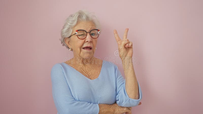 En glad äldre kvinna med grå hår med glasögon gör ett fredstecken mot en rosa bakgrund royaltyfri fotografi