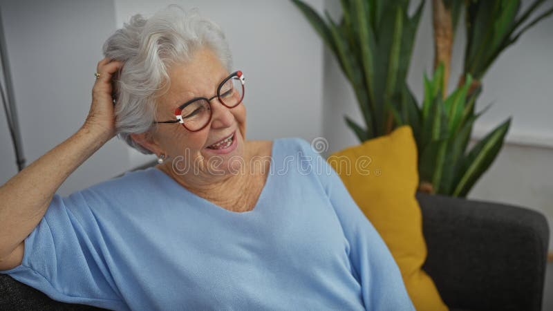 En glad äldre kvinna med grå hår med glasögon och blå övre relaxlar hemma på en soffa med gul kudde royaltyfri fotografi