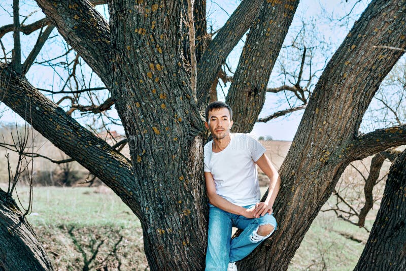 En grabb i en vit T-tröja och jeans sitter mellan träden royaltyfri bild En grabb i en vit T-tröja och jeans sitter mellan träden royaltyfri bild