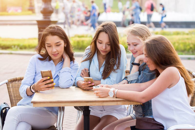 En grupp av ung flickastudenter som sitter på en tabell i parkera och med ett leende som diskuterar något som rymmer smartphones royaltyfria foton En grupp av ung flickastudenter som sitter på en tabell i parkera och med ett leende som diskuterar något som rymmer smartphones royaltyfria foton