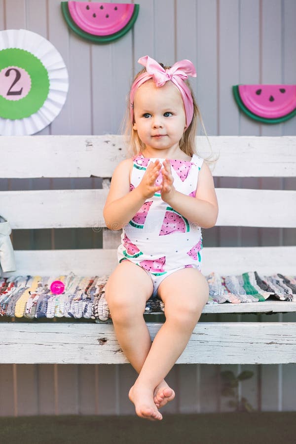 En gullig liten flicka i en jumpsuit sitter på en trävit bänk och applåderar henne händer royaltyfri bild En gullig liten flicka i en jumpsuit sitter på en trävit bänk och applåderar henne händer royaltyfri bild