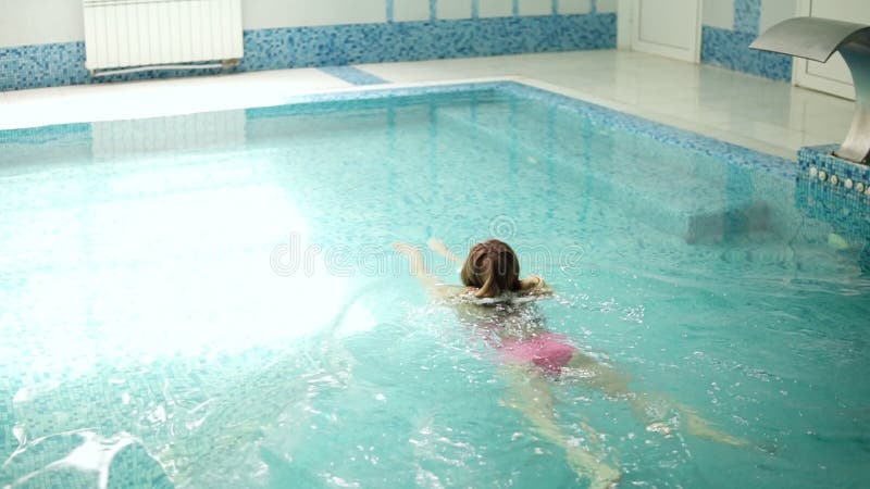 En härlig ung kvinna i en rosa baddräkt och ett långt hår simmar på den inomhus lilla pölen avkopplat långsamt