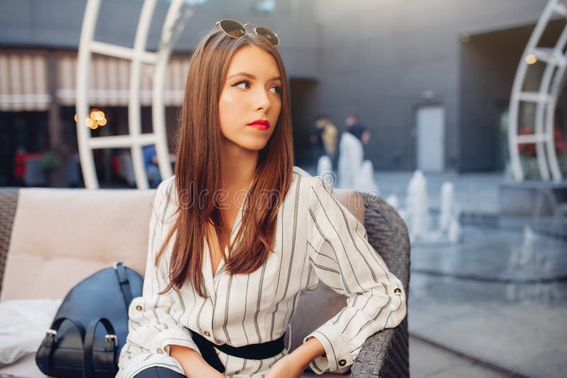 En iklädd ung härlig brunhårig flicka en randig trendig klänning sitter i ett utomhus- kafé för sommar och väntar på royaltyfri bild En iklädd ung härlig brunhårig flicka en randig trendig klänning sitter i ett utomhus- kafé för sommar och väntar på royaltyfri bild