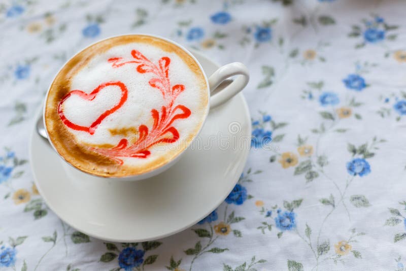 En kopp av varm cappuccino med en modell av en röd hjärta på tabellen med en bordduk i en blomma i en coffee shop Närbild och bl royaltyfri bild En kopp av varm cappuccino med en modell av en röd hjärta på tabellen med en bordduk i en blomma i en coffee shop Närbild och bl royaltyfri bild