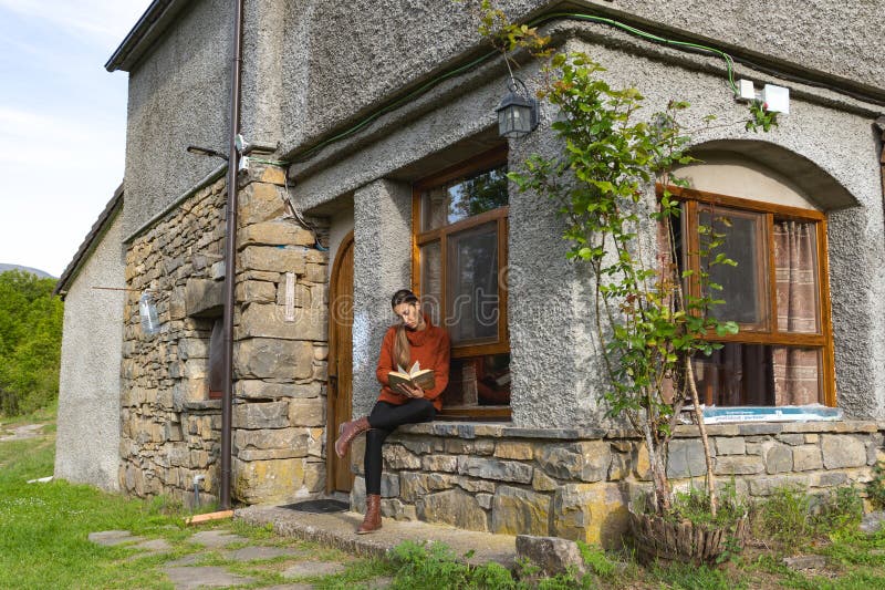 Woman with crossed legs reads at the entrance of a country house, concentrated, turns the page. Woman with crossed legs reads at the entrance of a country house, concentrated, turns the page