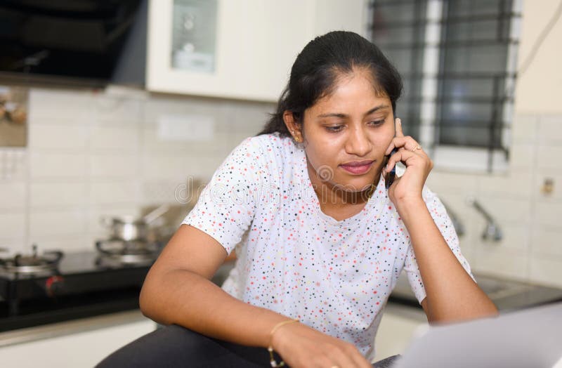 A determined Indian woman balances a phone call with reviewing documents in her makeshift home office kitchen. A determined Indian woman balances a phone call with reviewing documents in her makeshift home office kitchen