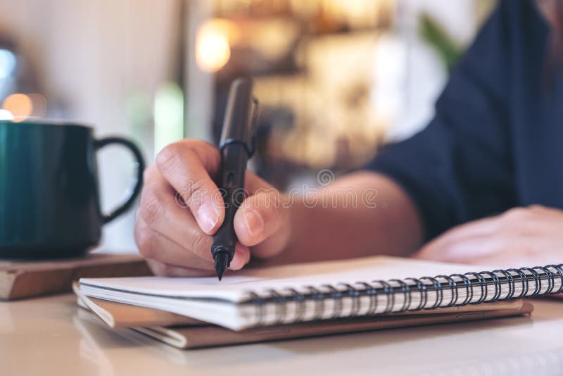 En kvinnas hand som skriver på den tomma anteckningsboken med kaffekoppen på tabellen i kafé royaltyfria foton En kvinnas hand som skriver på den tomma anteckningsboken med kaffekoppen på tabellen i kafé royaltyfria foton