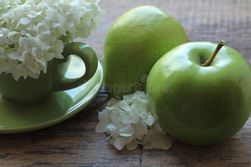 En la taza verde hay una inflorescencia magnífica de las flores blancas, y al lado de una pera verde con una manzana