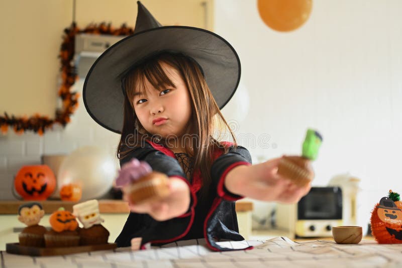 En lekfull flicka i häxkostumer som visar halloween cupcakes i köket royaltyfri fotografi