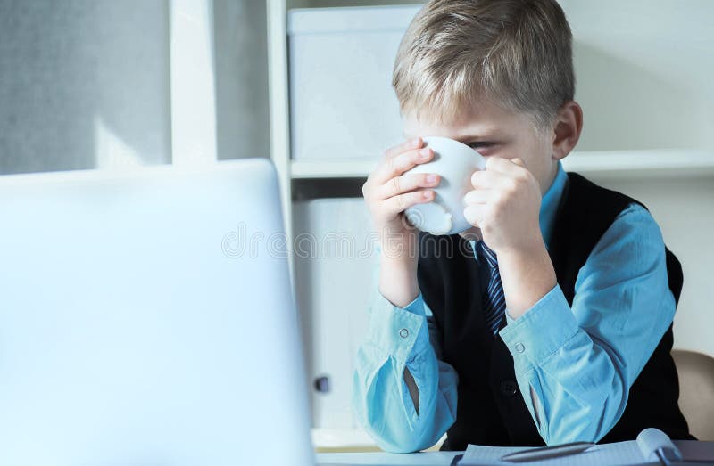 En liten affärsman som arbetar på en bärbar dator med en vit kopp kaffe eller varmt te Lustig liten chef på kontoret royaltyfria bilder