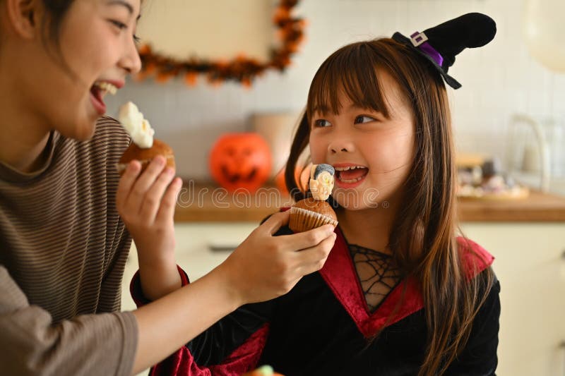 En liten flicka utklädd som häxa och mor gillar att äta halloween cupcakes i köket arkivbilder
