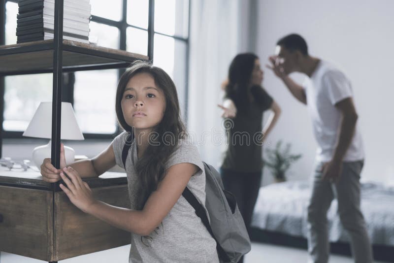 En liten mörker-haired flicka med en ryggsäck bak henne ser tillbaka bort, medan hon föräldrar grälar på henne bak henne tillbaka arkivfoto En liten mörker-haired flicka med en ryggsäck bak henne ser tillbaka bort, medan hon föräldrar grälar på henne bak henne tillbaka arkivfoto