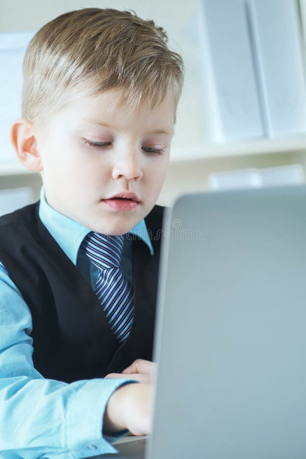 En liten söt pojke i affärskläder arbetar på datorn på kontoret och ler Småföretagare, liten chef royaltyfri bild