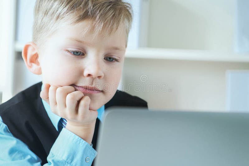 En liten söt pojke i affärskläder arbetar på datorn på kontoret och ler Småföretagare, liten chef royaltyfri bild
