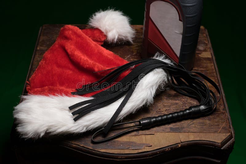 En luddig röd och vit jultomtenhatt, en svart piskar, och en flaska av motorisk olja, på ett gammalt wodden tabellen som fördelar arkivbilder En luddig röd och vit jultomtenhatt, en svart piskar, och en flaska av motorisk olja, på ett gammalt wodden tabellen som fördelar arkivbilder