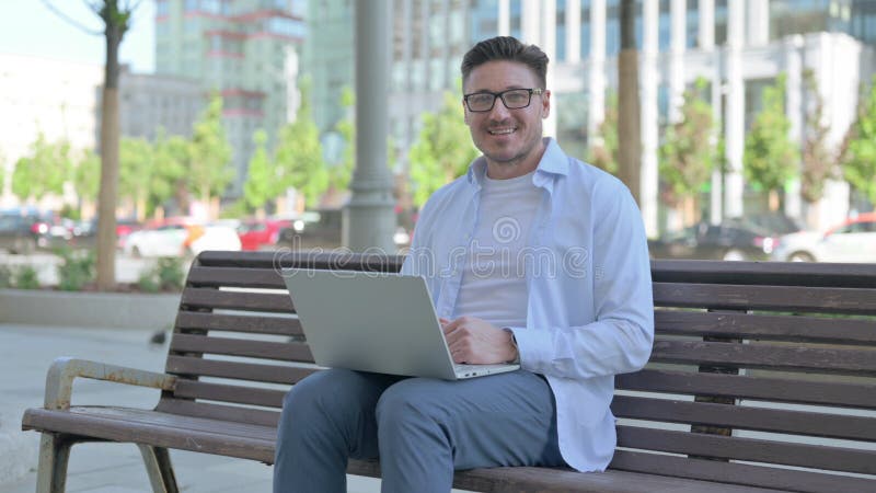 En man med bärbar dator som leker vid kameran medan man sitter utomhus på bänk arkivbilder
