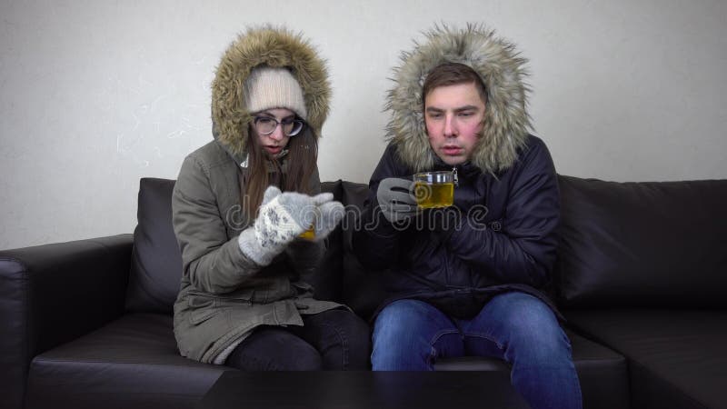 A men and a women sit at home in jackets and drink tea. The house is very cold because of the prohibition to raise the temperature in the house. A young couple sits without heating. 4k. A men and a women sit at home in jackets and drink tea. The house is very cold because of the prohibition to raise the temperature in the house. A young couple sits without heating. 4k