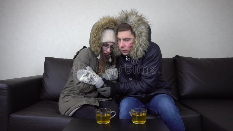 A men and a women sit at home in jackets and drink tea. The house is very cold because of the prohibition to raise the temperature in the house. A young couple sits without heating. 4k. A men and a women sit at home in jackets and drink tea. The house is very cold because of the prohibition to raise the temperature in the house. A young couple sits without heating. 4k