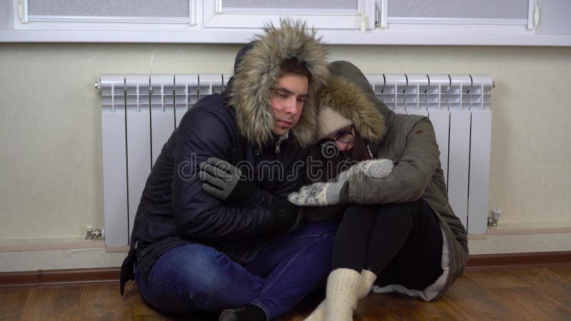 A man and a woman sit on the floor in their jackets by the radiator and warm themselves. A young married couple have not paid their bills and are freezing in the house. 4k. A man and a woman sit on the floor in their jackets by the radiator and warm themselves. A young married couple have not paid their bills and are freezing in the house. 4k