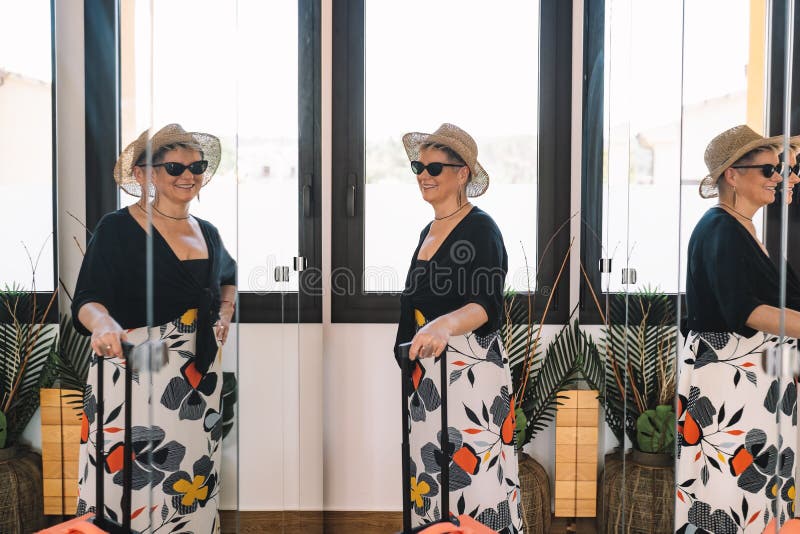 mature woman traveller, looking in the mirror while trying on her sunglasses for a walk along the coast on her summer holiday. bedroom, natural light, dressing room with mirrors. mature woman traveller, looking in the mirror while trying on her sunglasses for a walk along the coast on her summer holiday. bedroom, natural light, dressing room with mirrors.