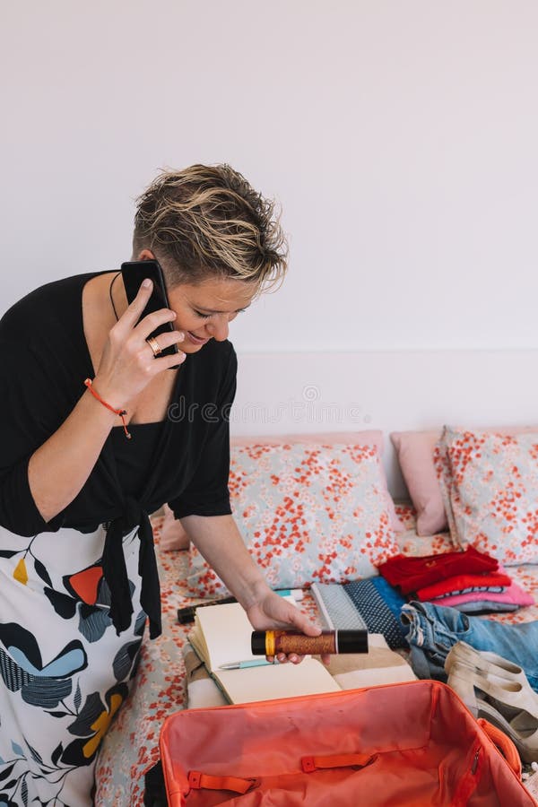 mature woman traveller using a smart phone and packing her suitcase preparing to travel on a summer holiday. Vertical, leisure and holiday concept. Room, illuminated by natural light, suitcase with clothes. mature woman traveller using a smart phone and packing her suitcase preparing to travel on a summer holiday. Vertical, leisure and holiday concept. Room, illuminated by natural light, suitcase with clothes.