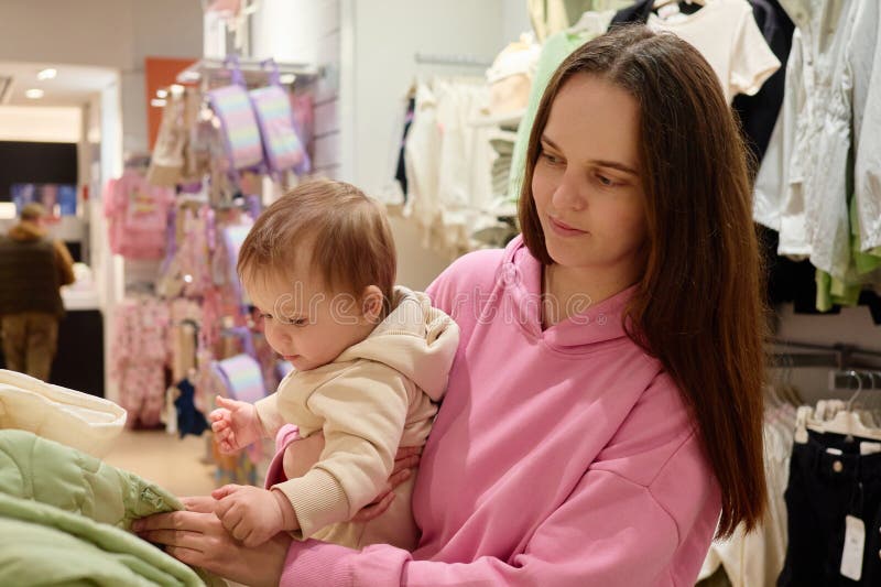 En mor håller barnet medan hon surfar på kläder i en butik. Barnet undersöker en väv som visar nyfikenhet i en varm inbjudan arkivbild