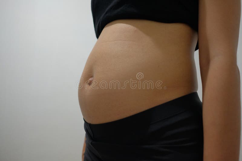 a 3 month old pregnant woman wearing a tank top and black pants is in front of a white wall. a 3 month old pregnant woman wearing a tank top and black pants is in front of a white wall