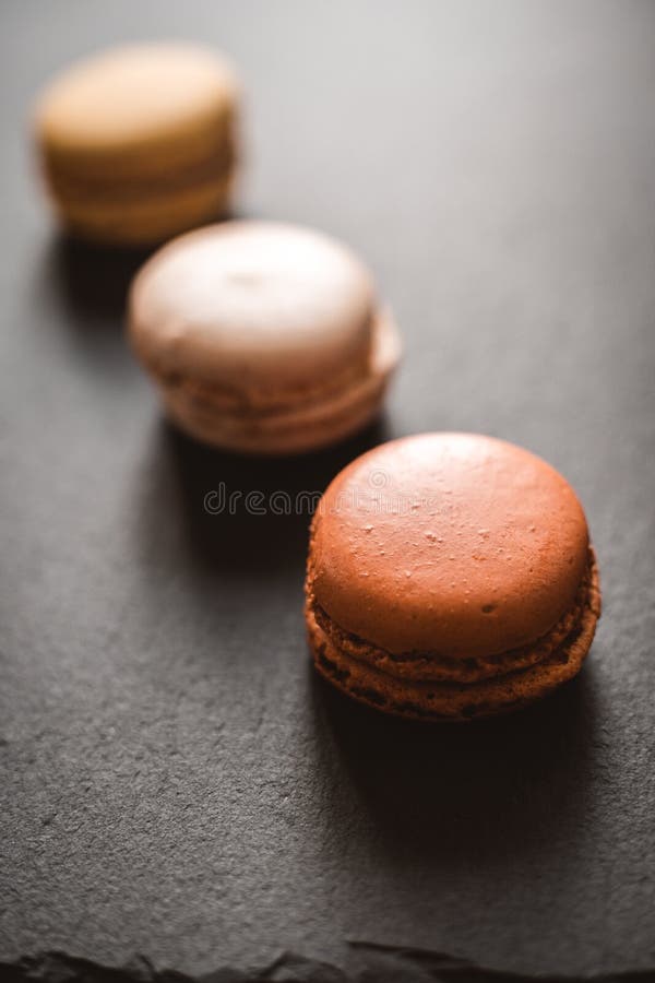 En mörk modig bild av chokladkaramel och vaniljmakaroner på en skifferbricka på en mörkgrå betongtextur royaltyfri foto