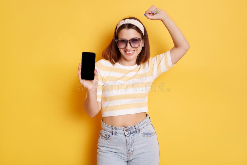 Positive satisfied woman wearing sunglasses, striped T-shirt and hair band, clenched fist, celebrating victory, betting and winning, showing phone with display, posing isolated over yellow background. Positive satisfied woman wearing sunglasses, striped T-shirt and hair band, clenched fist, celebrating victory, betting and winning, showing phone with display, posing isolated over yellow background