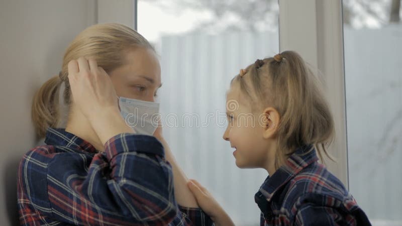 En omtänksam liten flicka som sitter på sängen placerar en medicinsk mask på mödrarnas ansikte lager videofilmer