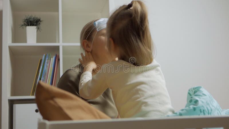 En omtänksam liten flicka som sitter på sängen placerar en medicinsk mask på mödrarnas ansikte arkivfilmer