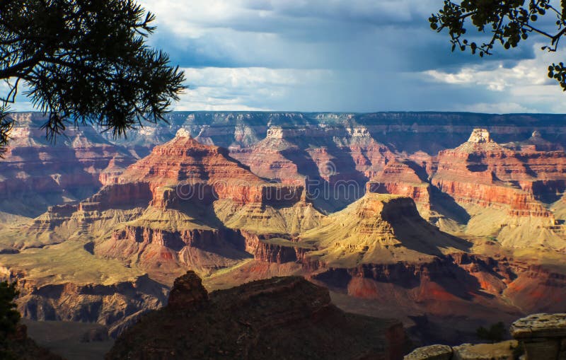 En sikt av Grand Canyon med vaggar bildande som markeras av solen på en mulen dag som inramas av träd i förgrunden royaltyfria bilder En sikt av Grand Canyon med vaggar bildande som markeras av solen på en mulen dag som inramas av träd i förgrunden royaltyfria bilder