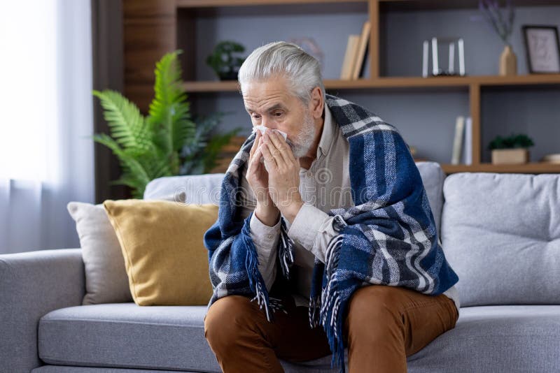 A sick gray-haired man is sitting at home on the sofa covered with a warm blanket, blowing his nose from a cold, suffering from colds and the flu. A sick gray-haired man is sitting at home on the sofa covered with a warm blanket, blowing his nose from a cold, suffering from colds and the flu.
